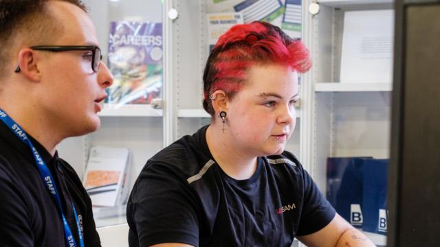 Young person with red hair sat with staff member. They are using a computer.